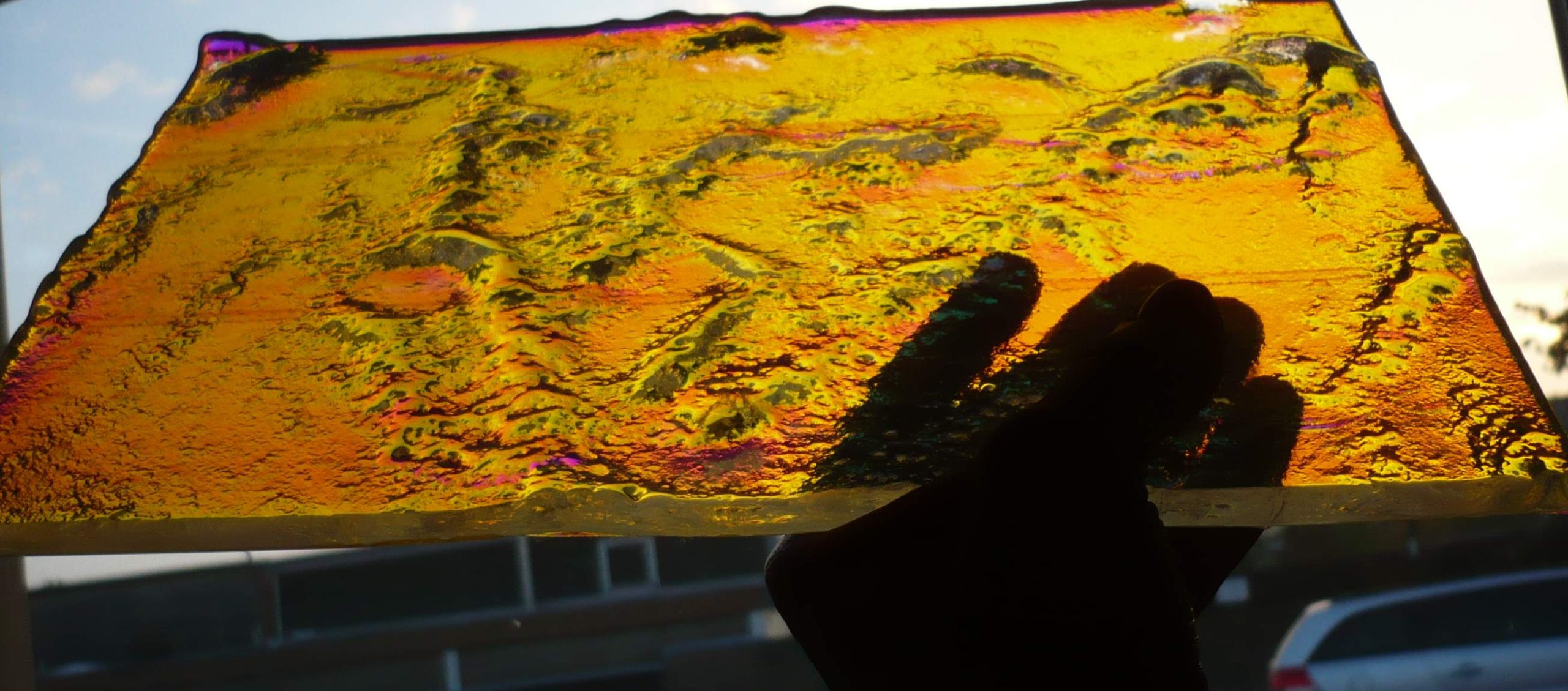 Hand haelt skulpturales Schmelzglas-Stueck in leuchtendem Bernstein-Orange gegen das Licht mit bewegtem Relief