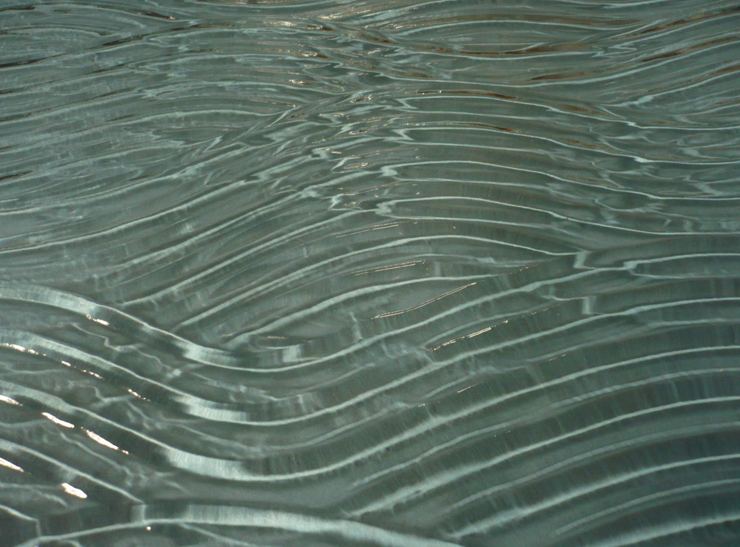 Klare Schmelzglas-Platte mit sanftem horizontalem Wellenrelief und fliessender Oberflaechenstruktur in Nahansicht